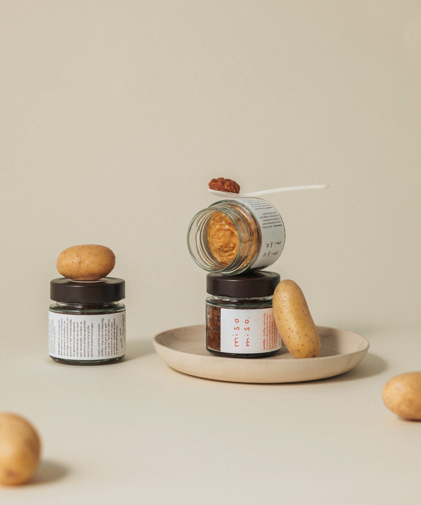 ästhetisches stillleben von miso miso gläsern auf beigem hintergrund. ein geöffnetes glas liegt schräg auf einem anderen, daneben und darauf sind frische kartoffeln arrangiert. ein kleiner löffel mit dunkler miso-paste zeigt die fertige würzpaste.