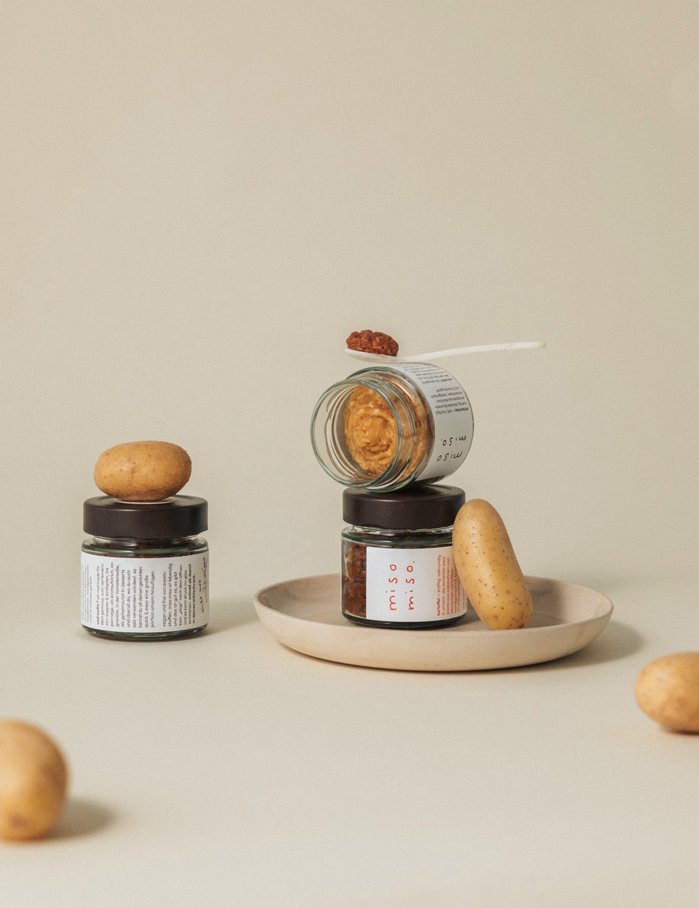 ästhetisches stillleben von miso miso gläsern auf beigem hintergrund. ein geöffnetes glas liegt schräg auf einem anderen, daneben und darauf sind frische kartoffeln arrangiert. ein kleiner löffel mit dunkler miso-paste zeigt die fertige würzpaste.