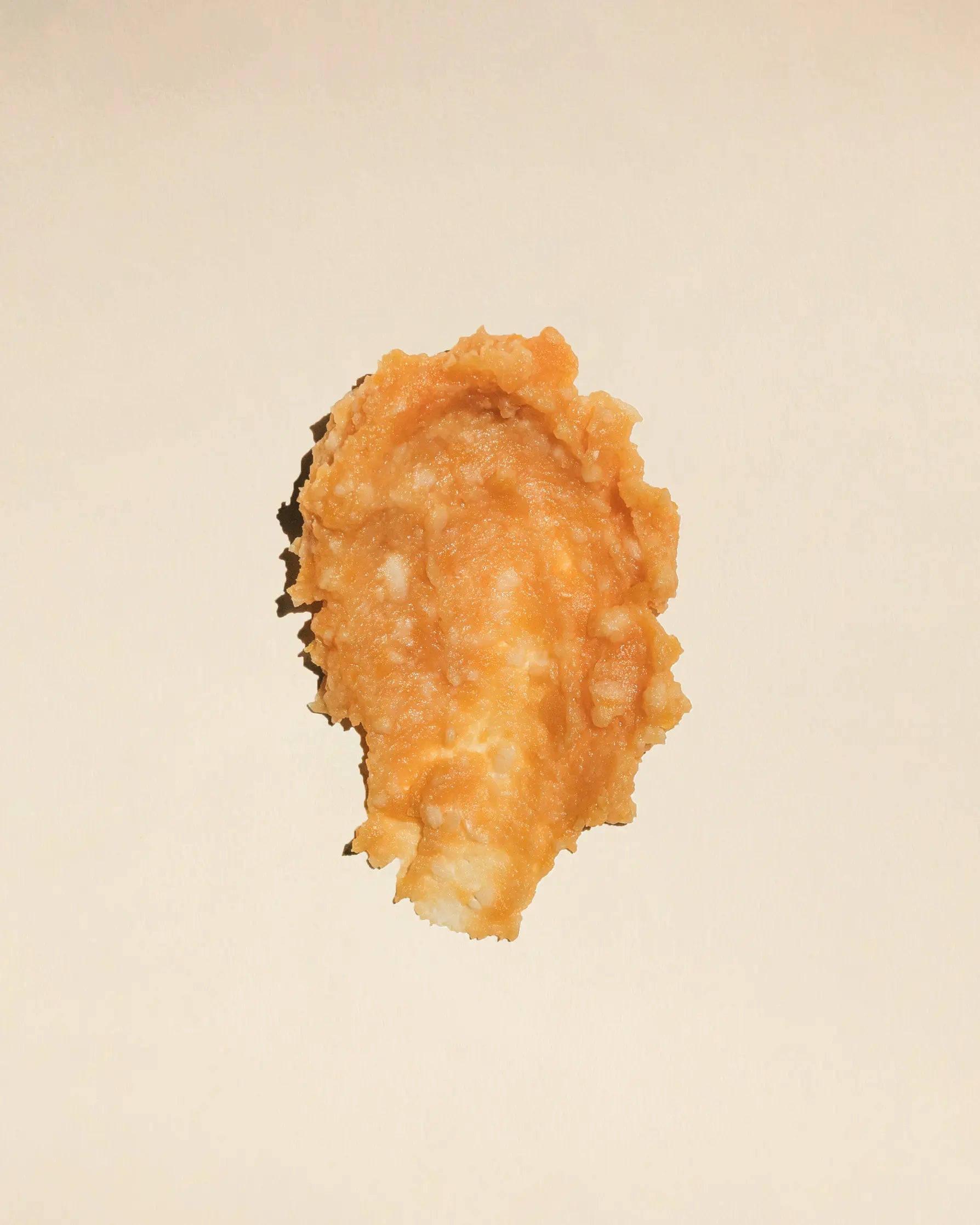 detailaufnahme einer cremigen kichererbsen-miso-paste auf hellem, beigen hintergrund. die makro-ansicht zeigt die körnige, natürliche textur und die warmen gold-orange-töne der unpasteurisierten fermentation.