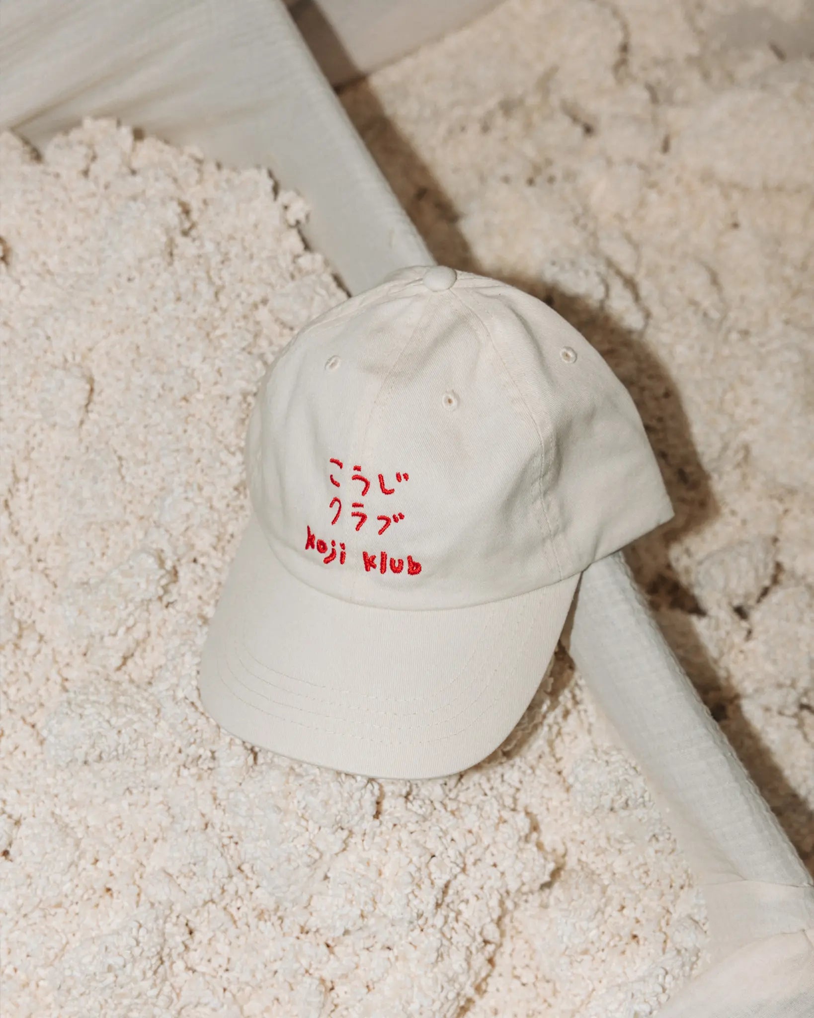 Weiße Koji Klub Cap mit rotem Stick-Logo über einer Wanne mit fermentiertem Koji-Reis
