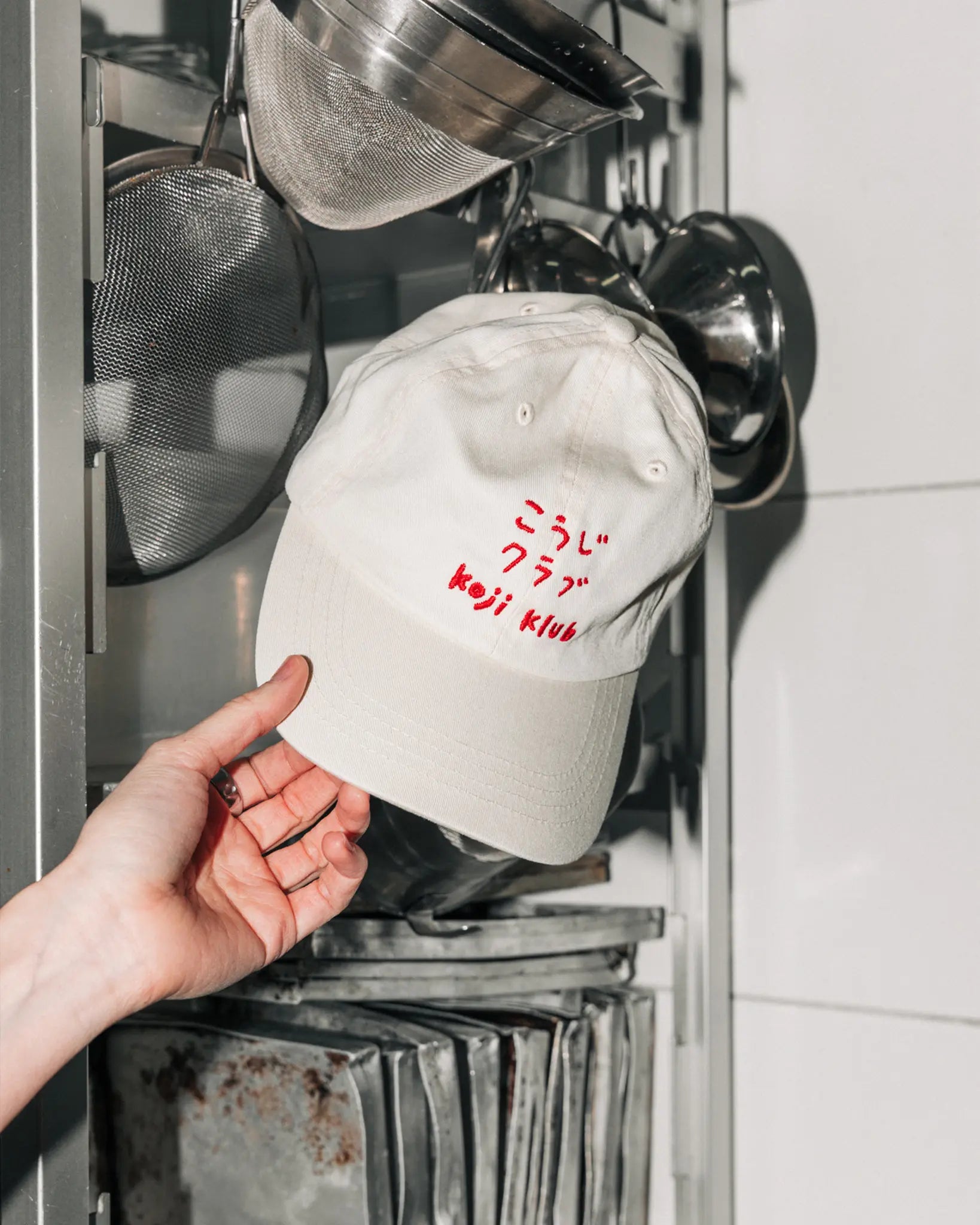 Weiße Koji Klub Cap mit rotem Stick-Logo an einem Metallregal in der Kueche.