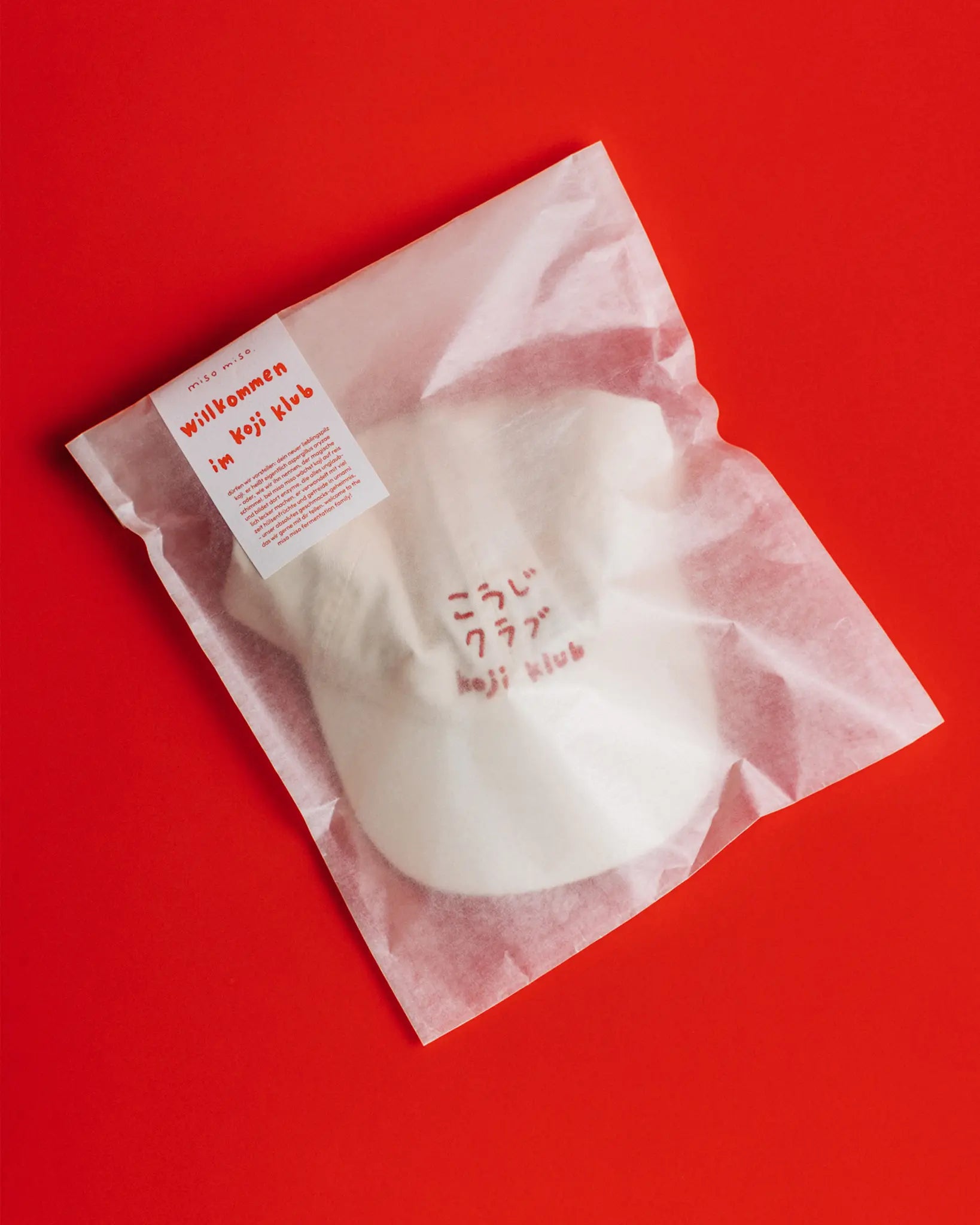 Weiße Koji Klub Cap mit rotem Stick-Logo in Verpackung auf rotem Hintergrund.