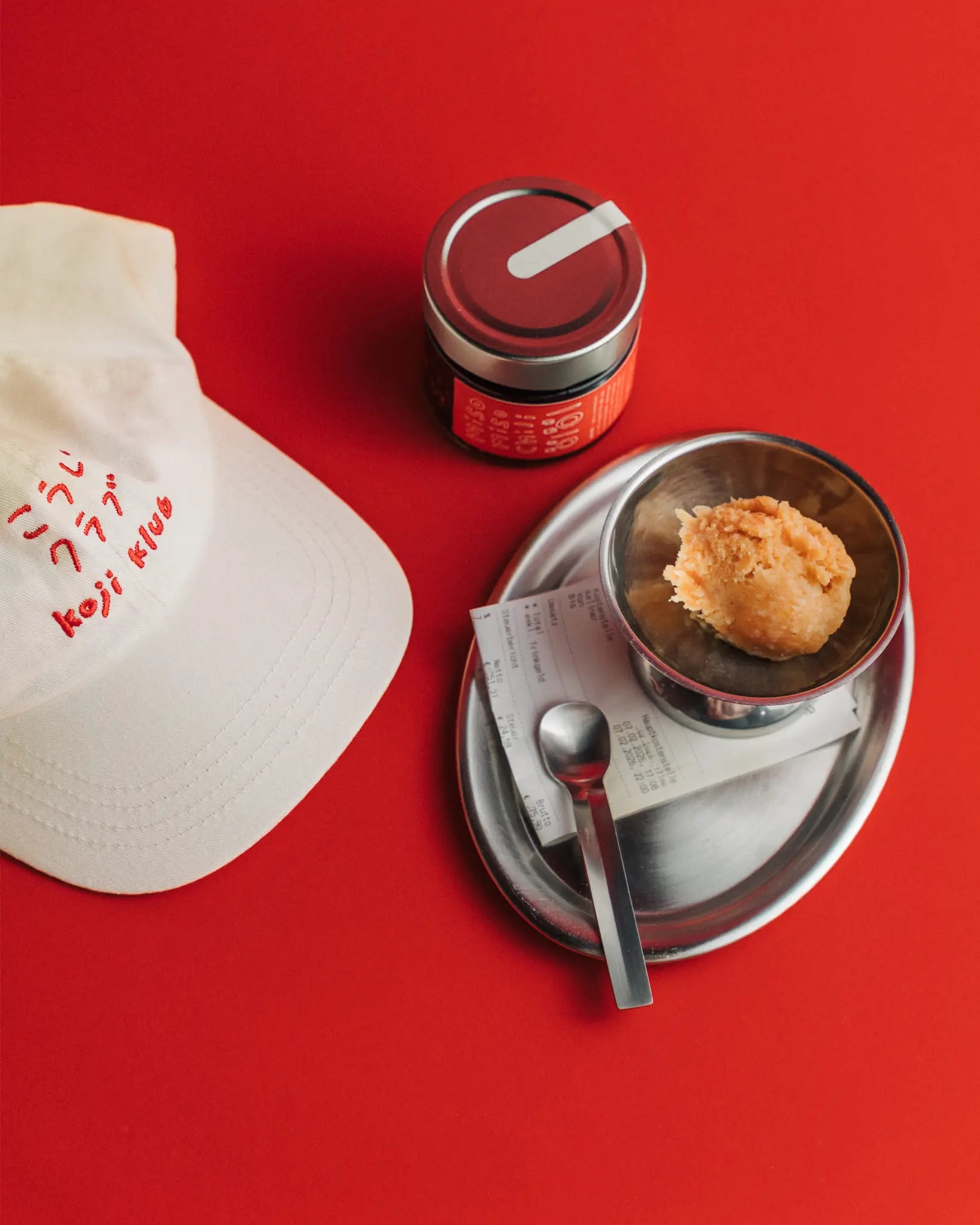 Weiße Koji Klub Cap mit rotem Stick-Logo und Kichererbsen miso miso und Tablett auf rotem Hintergrund