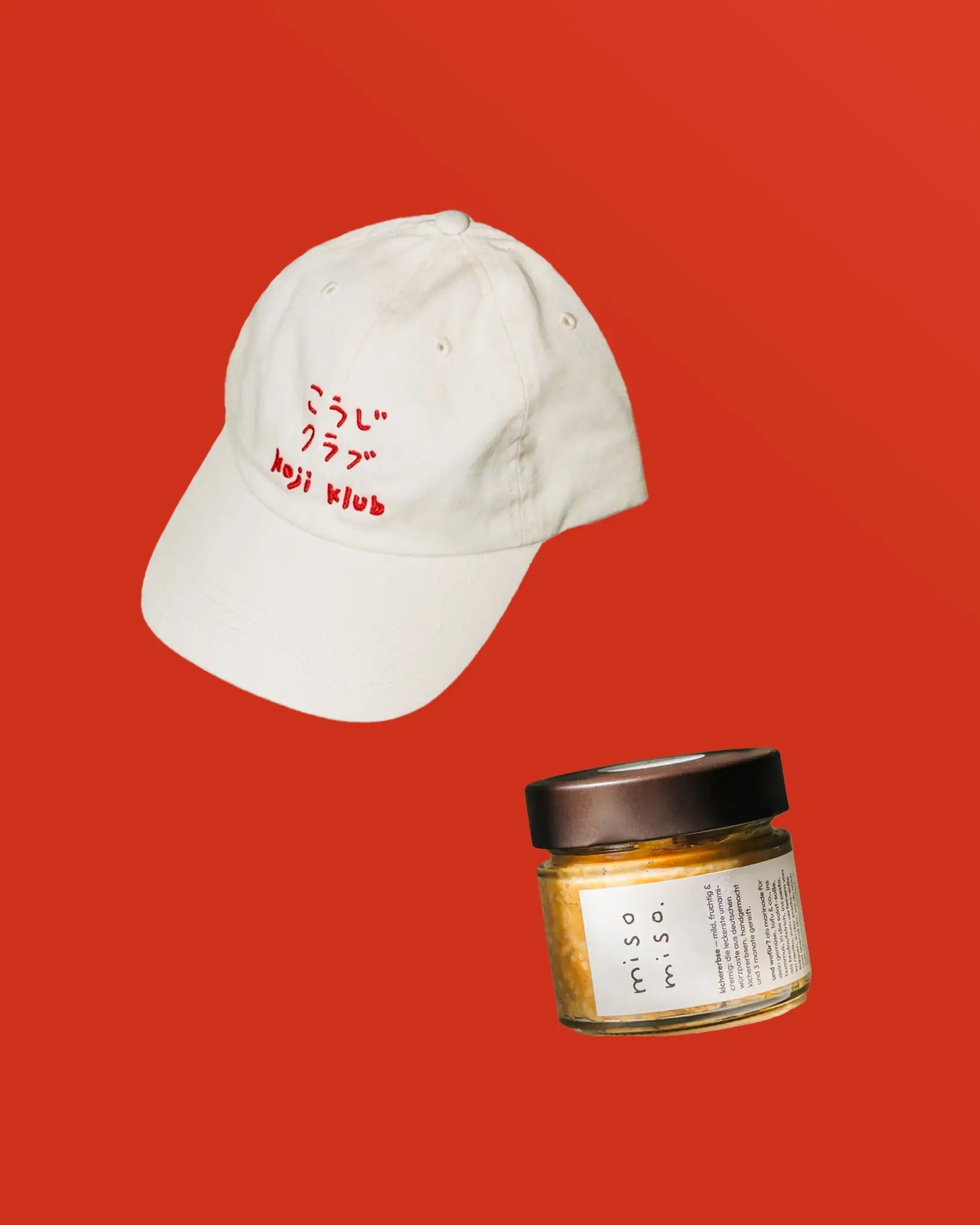 Weiße Koji Klub Cap mit rotem Stick-Logo und Kichererbsen miso miso als Bundle auf rotem Hintergrund